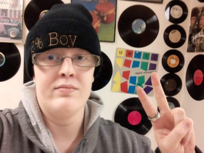 Veld looks into the camera, holding up the peace sign in front of a wall of vinyl records.